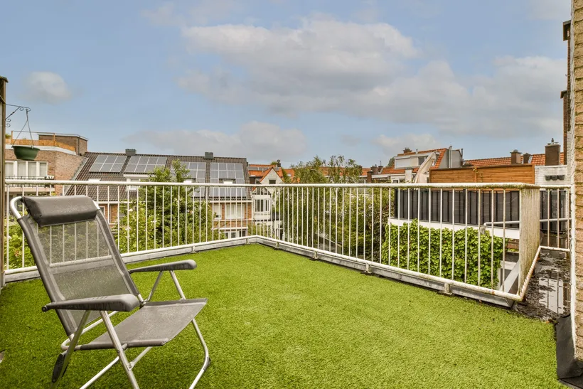 A roof terrace with artificial grass laid up to the railing. At the right-hand edge, where the grass ends, the dark roof surface shows, with puddles and dirt collected along it.
