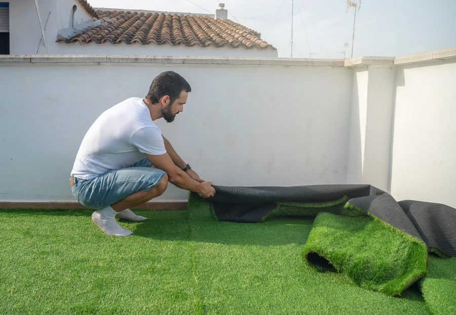 Un hombre en cuclillas tiende un rollo de césped artificial sobre una azotea de muros encalados, con un tejado de teja árabe y antenas detrás. El césped ya cubre el suelo y el rollo deja ver el soporte negro.