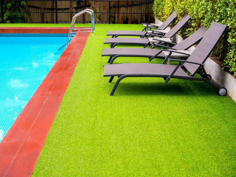 Artificial grass running right up to the red stone edge of a pool, with four grey sun loungers on it and a hedge behind.