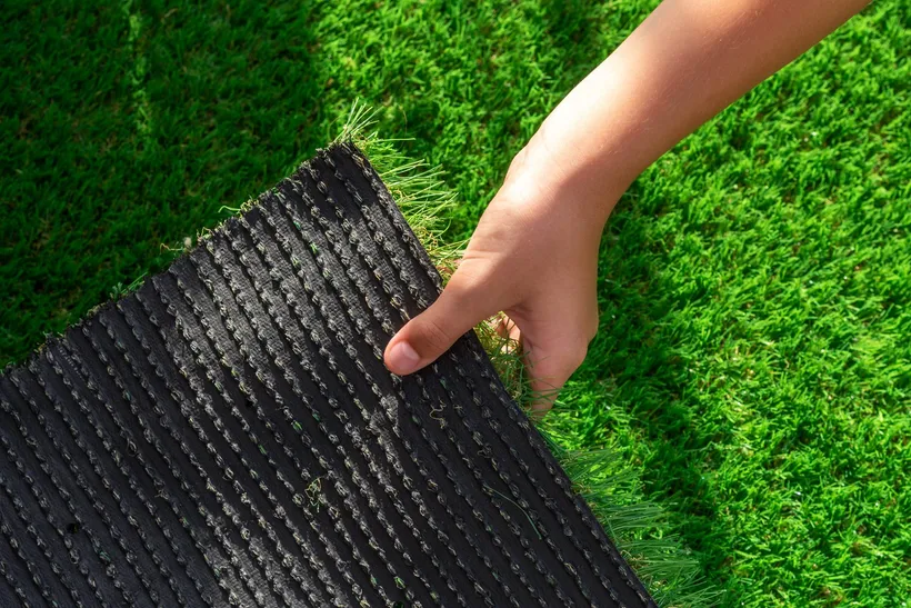 A hand lifting a corner of artificial grass to show the backing underneath: black fabric with rows of stitches holding the fibres, no holes visible.