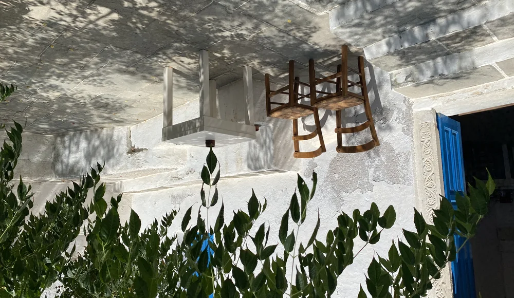 Mesa y dos sillas contra un muro encalado, con toda la solera cubierta por la sombra moteada de la copa de un árbol.