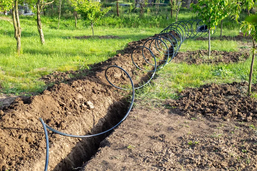 Zanja larga abierta en un terreno con hierba, con la tierra amontonada a un lado y una tubería negra de riego enrollada a lo largo.